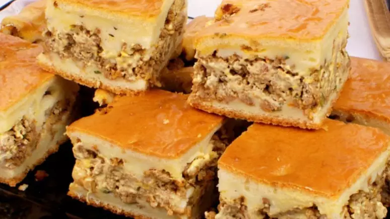 Várias fatias de torta sobre um recipiente, cortadas em formatos quadrados, seus recheios estão à mostra, sendo eles de carne moída.
